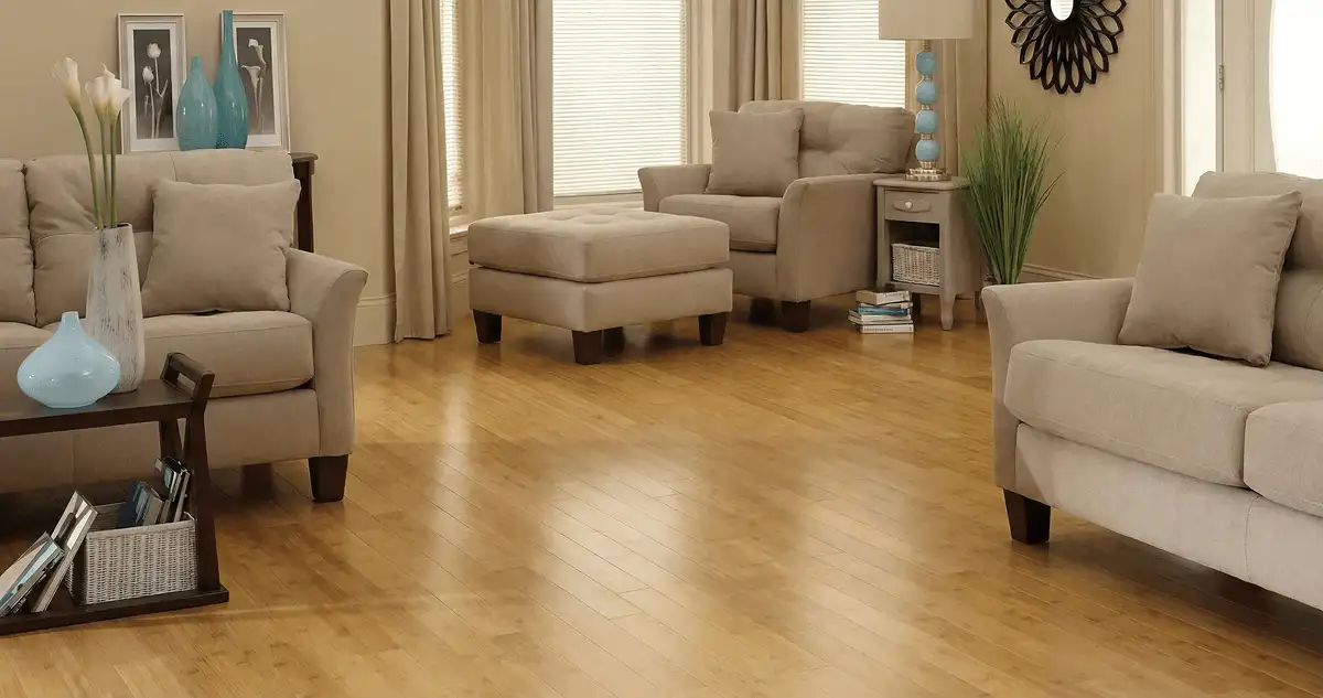 A living room with professionally installed bamboo flooring, beige sofas, a matching ottoman, a side table with a blue lamp, vases, framed pictures, and large windows with blinds and curtains in Arlington, TX